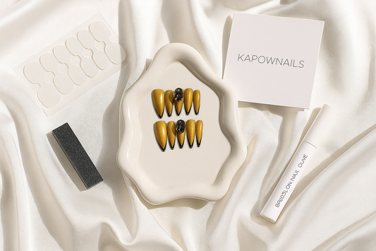 Elegant flat lay of glossy press-on nails, nail glue, buffer, and adhesive tabs arranged on a white silk cloth with soft daylight and shadows. Include a minimal KAPOWNAILS logo card in the corner. Feminine, premium, modern aesthetic.
