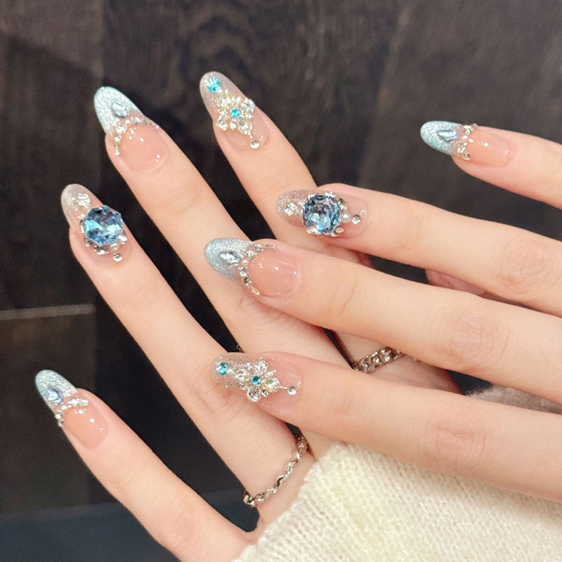 Coastal-inspired almond nails with aqua-silver sparkle, ornate rhinestone patterns & geode centerpiece - wearable luxury art